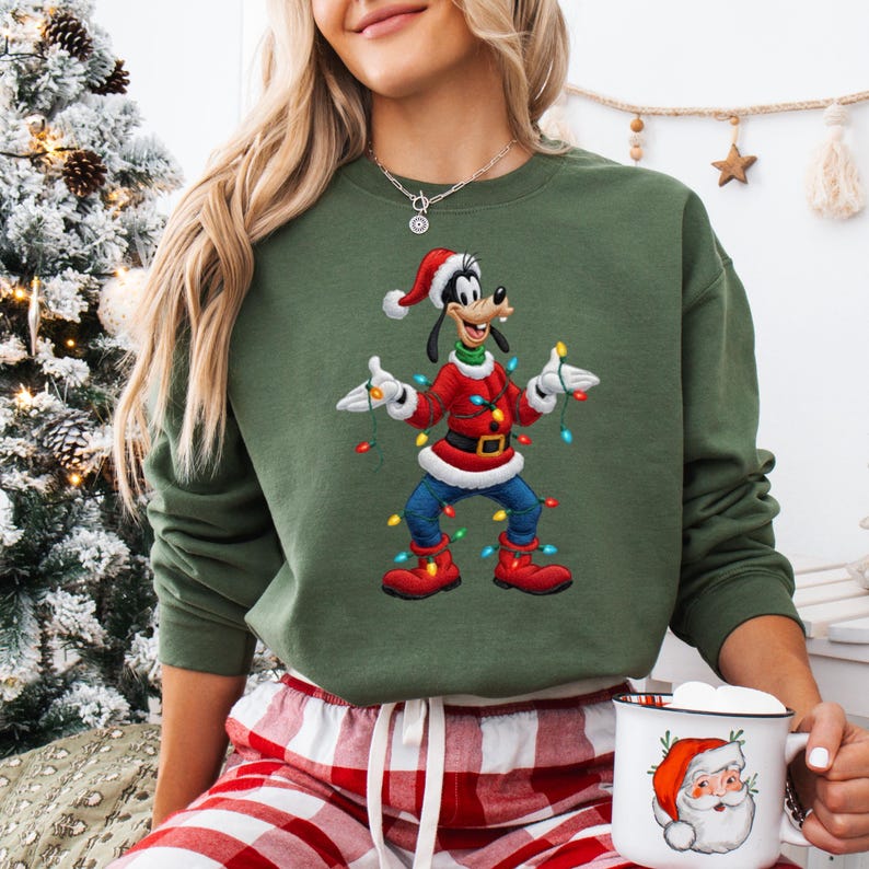 May include: An olive green sweatshirt with a cartoon Goofy character in a Santa hat and red Christmas attire, holding festive lights. The sweatshirt is worn with red and white plaid pyjama bottoms. A white mug with a Santa Claus design is also present.