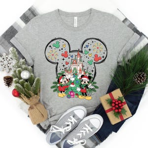 Puede incluir: Una camiseta gris con un dise&ntilde;o de silueta de Mickey Mouse. La silueta presenta a Mickey y Minnie Mouse decorando un &aacute;rbol de Navidad frente al Castillo de Cenicienta. El dise&ntilde;o incluye fuegos artificiales y corazones.
