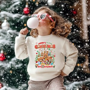 May include: A child wearing a cream-colored sweatshirt with a colorful graphic featuring Disney characters and the text "Mickey's Very Merry Christmas" and "Walt Disney World". The child is wearing pink sunglasses and blowing a bubble.