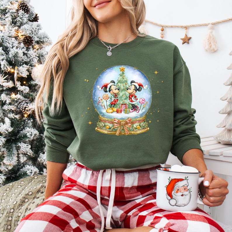 May include: Green sweatshirt with a snow globe design featuring Mickey and Minnie Mouse, a Christmas tree, and presents. The snow globe is set against a starry background. The sweatshirt is a long-sleeved crew neck.