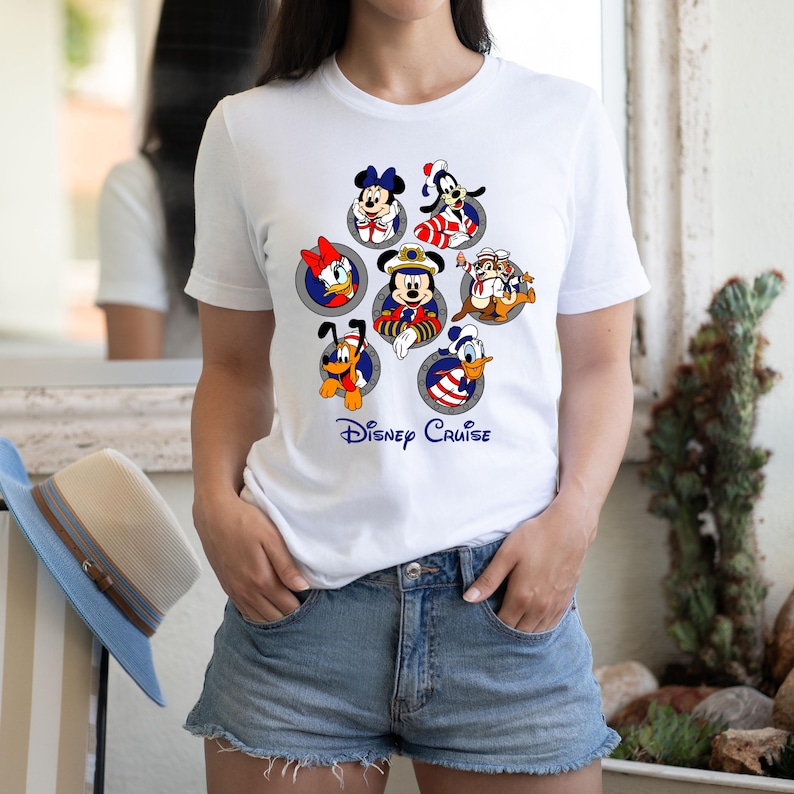 May include: White t-shirt featuring a colorful graphic of Disney characters, including Mickey Mouse, Minnie Mouse, Donald Duck, and Goofy, in circular frames. The words "Disney Cruise" are printed in blue script below the characters. The shirt is paired with denim shorts.