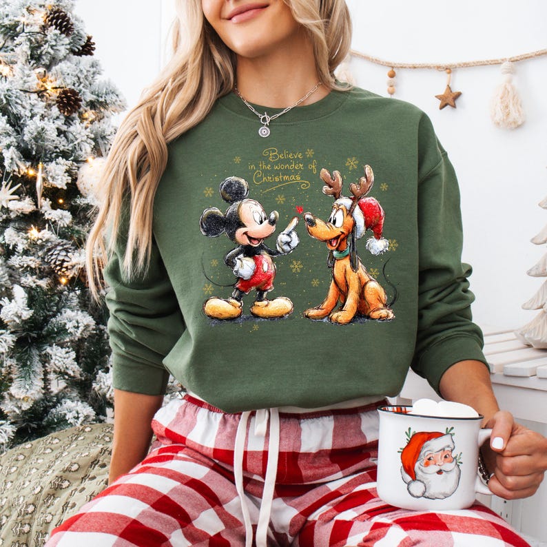 May include: Green sweatshirt featuring Mickey Mouse and Pluto with reindeer antlers, and the text "Believe in the wonder of Christmas." The sweatshirt is paired with red and white plaid pajama pants. A mug with a Santa Claus illustration is also visible.