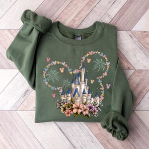 May include: An olive green sweatshirt featuring a castle design with a Mickey Mouse head outline made of hearts and fireworks. The castle has blue turrets and is surrounded by flowers. The sleeves are cuffed.