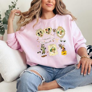 May include: A light pink sweatshirt featuring various Disney character graphics. The design includes Mickey and Minnie Mouse, Donald Duck, and Goofy, with the words "ANIMAL KINGDOM" in a decorative font. The sweatshirt has long sleeves.
