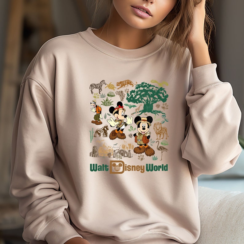 May include: A tan sweatshirt featuring a graphic print of Mickey Mouse and Goofy dressed in safari outfits. The print also includes a variety of animals, plants, and the text "Walt Disney World".