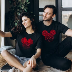 May include: A couple wearing matching black t-shirts with red heart-shaped Mickey Mouse and Minnie Mouse designs. The woman is wearing a red bow on her Minnie Mouse design.