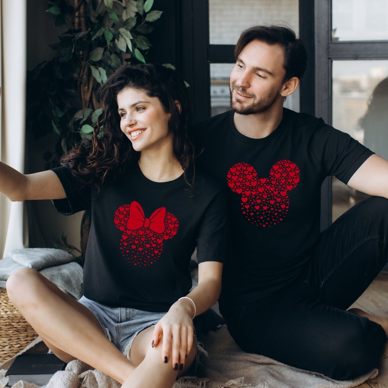 May include: A couple wearing matching black t-shirts with red heart-shaped Mickey Mouse and Minnie Mouse designs. The woman is wearing a red bow on her Minnie Mouse design.