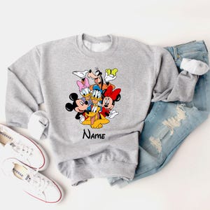 Op de afbeelding: Grijs sweatshirt met een kleurrijke afbeelding van Mickey Mouse, Minnie Mouse, Donald Duck, Goofy en Pluto. De tekst "Name" staat onder de afbeelding.