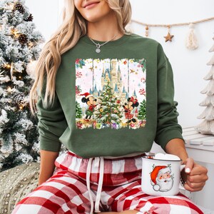 Może przedstawiać: Zielona bluza z świąteczną grafiką Myszki Miki i Myszki Minnie dekorujących choinkę przed zamkiem. Bluza jest połączona z czerwono-białymi spodniami od piżamy w kratę. W dłoni trzymany jest biały kubek z motywem Świętego Mikołaja.