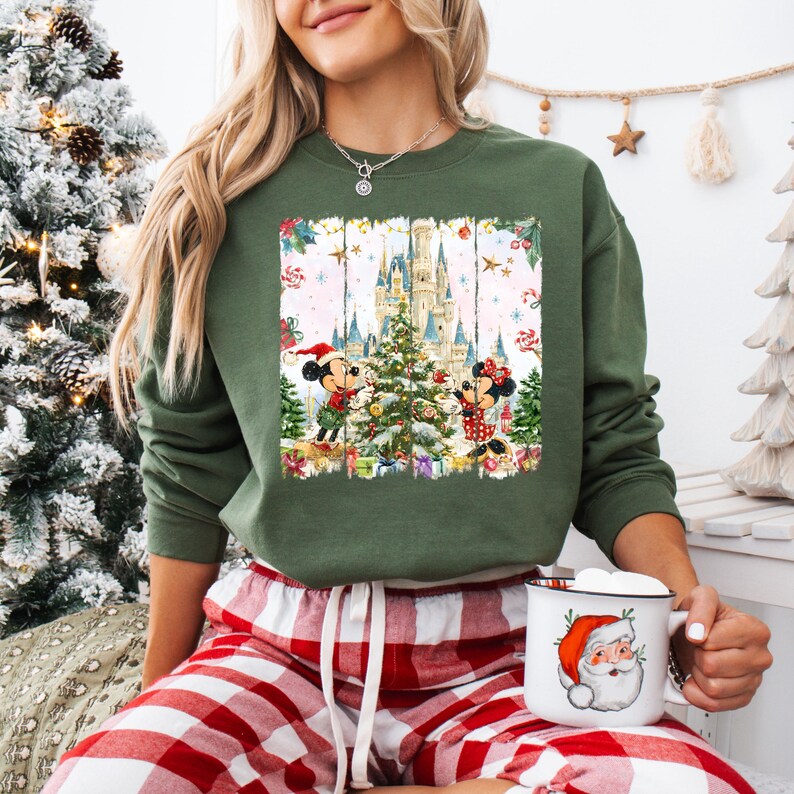 Może przedstawiać: Zielona bluza z świąteczną grafiką Myszki Miki i Myszki Minnie dekorujących choinkę przed zamkiem. Bluza jest połączona z czerwono-białymi spodniami od piżamy w kratę. W dłoni trzymany jest biały kubek z motywem Świętego Mikołaja.
