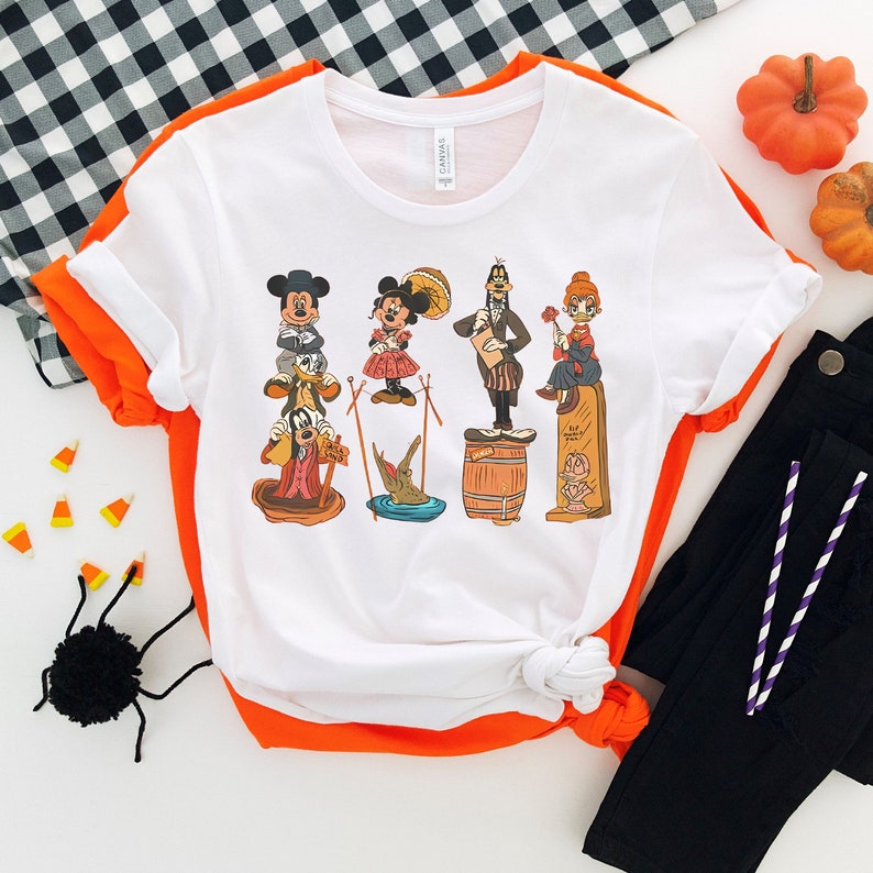 May include: White t-shirt featuring a colourful cartoon image of Mickey Mouse, Minnie Mouse, Goofy, Donald Duck, and Pluto. The characters are dressed in Halloween costumes.