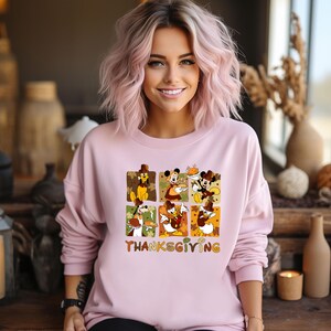 May include: A light pink sweatshirt featuring a Thanksgiving-themed graphic with Disney characters. The graphic includes images of Mickey Mouse, Minnie Mouse, Donald Duck, and Goofy in autumnal settings. The word "Thanksgiving" is printed in a colorful font.