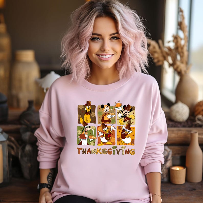 May include: A light pink sweatshirt featuring a Thanksgiving-themed graphic with Disney characters. The graphic includes images of Mickey Mouse, Minnie Mouse, Donald Duck, and Goofy in autumnal settings. The word "Thanksgiving" is printed in a colorful font.