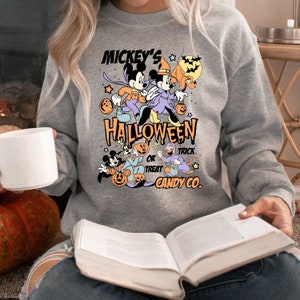 May include: A gray sweatshirt with a Mickey Mouse Halloween design. The design features Mickey and Minnie Mouse dressed in Halloween costumes, along with Pluto and Goofy. The text on the sweatshirt reads "Mickey's Halloween Trick or Treat Candy Co."