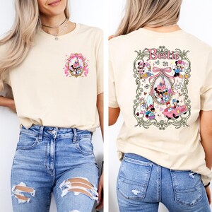 May include: A beige t-shirt with a pink and gold design featuring Mickey Mouse and Minnie Mouse on the front and a larger design on the back with the text "Bestie" and Mickey Mouse and Minnie Mouse characters in a car and other scenes.