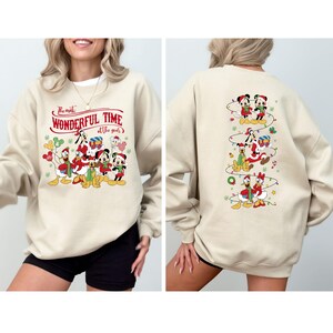 May include: A beige sweatshirt featuring a festive design with Disney characters. The front displays Mickey, Minnie, Donald, and Goofy with the text "The most Wonderful Time of the year". The back has a vertical design of characters with Christmas lights.