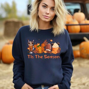 K&ouml;nnte beinhalten: Ein marineblauer Pullover mit einem Aufdruck einer K&uuml;rbis, einem Football und einer Tasse hei&szlig;er Schokolade. Der Text "Tis The Season" ist unter dem Aufdruck gedruckt.