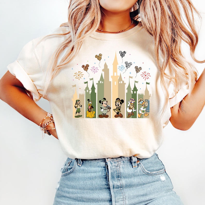 May include: A white t-shirt with a graphic of a castle, fireworks, and Disney characters. The characters are Mickey Mouse, Minnie Mouse, Donald Duck, Goofy, Pluto, and Chip and Dale. The castle is in shades of green and brown. The fireworks are red, yellow, and blue. The hearts are in various colors and patterns.