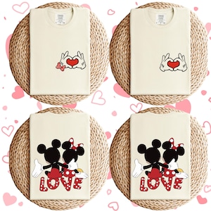 May include: Set of four cream-colored t-shirts with a Mickey Mouse and Minnie Mouse design. Two shirts feature Mickey and Minnie holding hands with a red heart in the middle. The other two shirts feature Mickey and Minnie holding hands with the word "LOVE" in red letters.