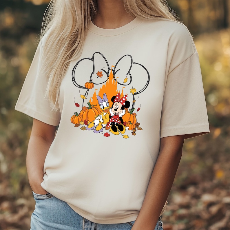 May include: Beige t-shirt featuring a graphic of Minnie Mouse and Daisy Duck in front of a castle with pumpkins and fall leaves. Minnie Mouse is wearing a red polka dot bow and dress. Daisy Duck is wearing a purple bow and dress.