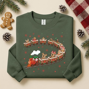 May include: Green sweatshirt featuring a festive train design. The train is red and green, with a white smoke cloud. The train cars are filled with gingerbread men and cartoon characters. Red and green snowflakes are scattered around the train.