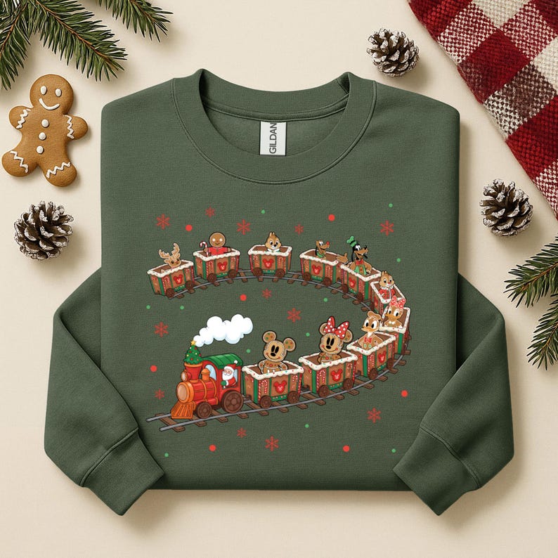 May include: Green sweatshirt featuring a festive train design. The train is red and green, with a white smoke cloud. The train cars are filled with gingerbread men and cartoon characters. Red and green snowflakes are scattered around the train.