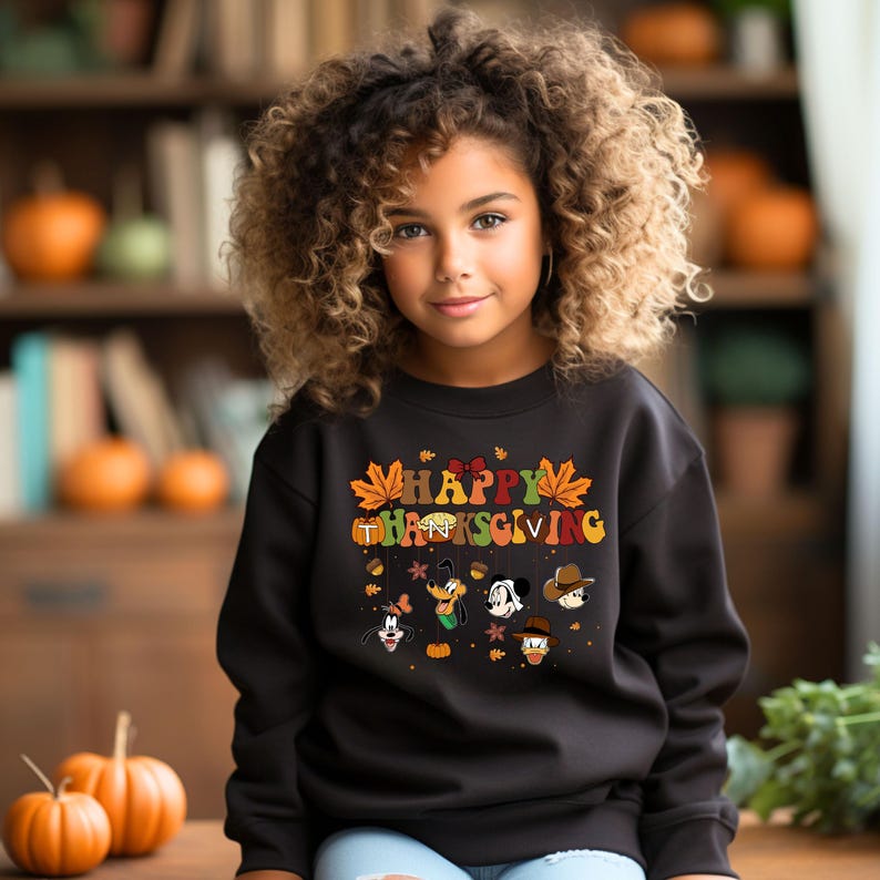 Op de afbeelding: Een jong persoon draagt een zwarte trui met de woorden "HAPPY THANKSGIVING" in kleurrijke letters en herfstthema-afbeeldingen. De trui heeft stripfiguren en herfstbladeren. Pompoenen staan op de voorgrond.