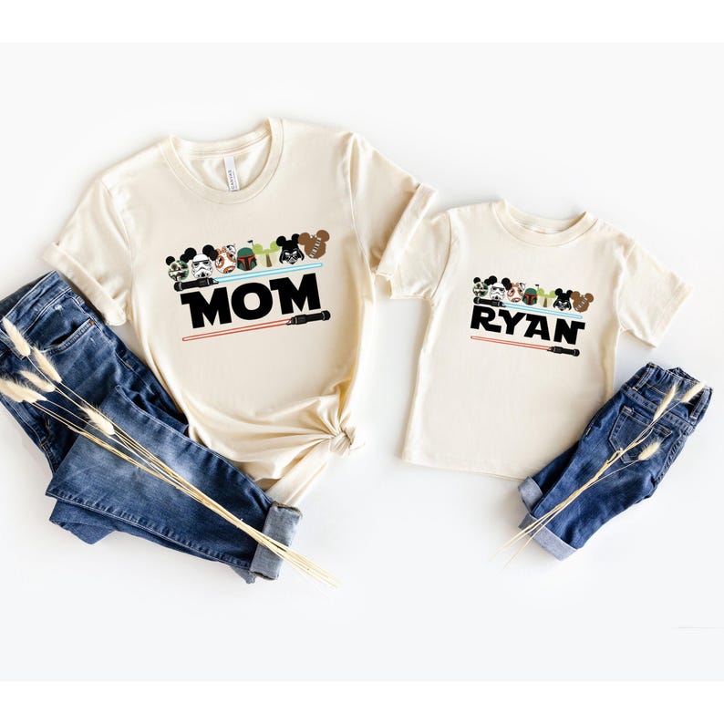 May include: Two cream-colored t-shirts with Star Wars-themed graphics. One shirt says "MOM" and the other says "RYAN" in black block letters. Both shirts have a row of cartoon characters and a red lightsaber design. Blue jeans are next to the shirts.