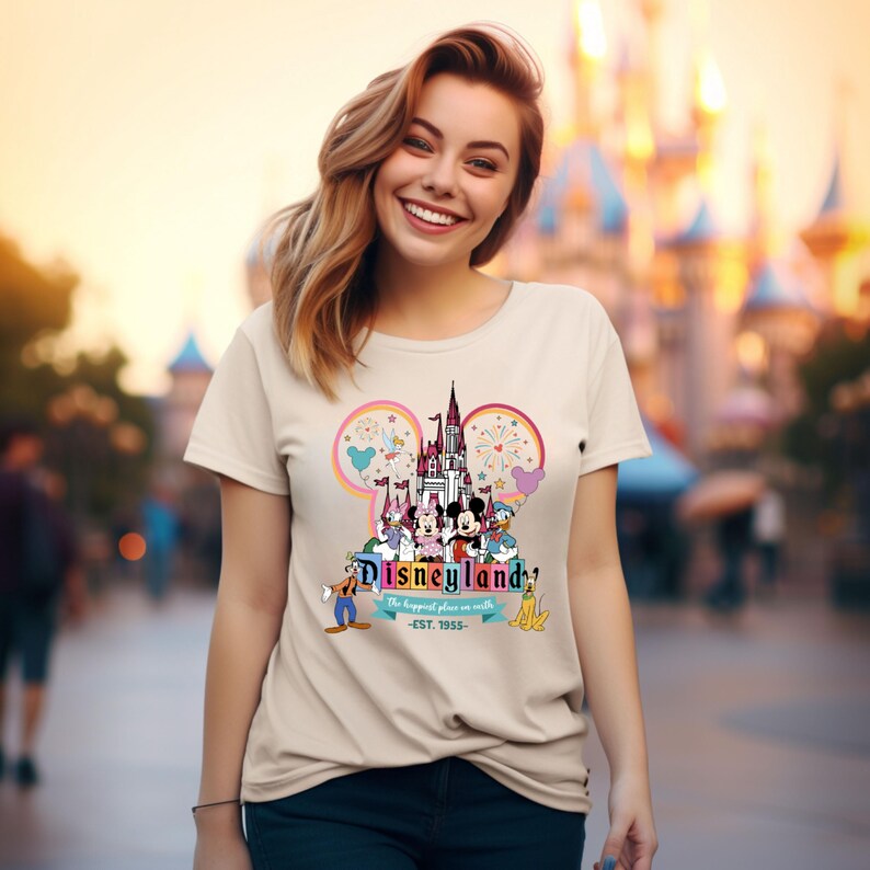 May include: A beige t-shirt with a colorful graphic of Mickey Mouse and friends in front of the Disneyland castle. The text "Disneyland" is printed in a colorful font with the tagline "The happiest place on earth - EST. 1955 -" printed below.