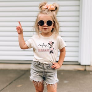 K&ouml;nnte beinhalten: Ein junges Kind tr&auml;gt ein cremefarbenes T-Shirt mit dem Text "I'm this Minnie" und einer Minnie Mouse-Grafik. Das Kind tr&auml;gt auch graue Jeansshorts und eine runde Sonnenbrille. Das Kind zeigt mit einem Finger nach oben.