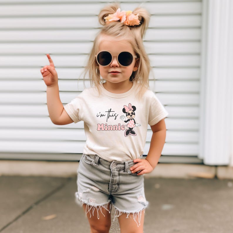 K&ouml;nnte beinhalten: Ein junges Kind tr&auml;gt ein cremefarbenes T-Shirt mit dem Text "I'm this Minnie" und einer Minnie Mouse-Grafik. Das Kind tr&auml;gt auch graue Jeansshorts und eine runde Sonnenbrille. Das Kind zeigt mit einem Finger nach oben.
