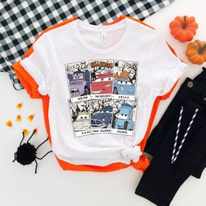 May include: White t-shirt with a Halloween themed graphic featuring characters from the Cars movie franchise. The graphic shows Mater, McQueen, Sally, Luigi, Doc Hudson, and Guido in a spooky setting.