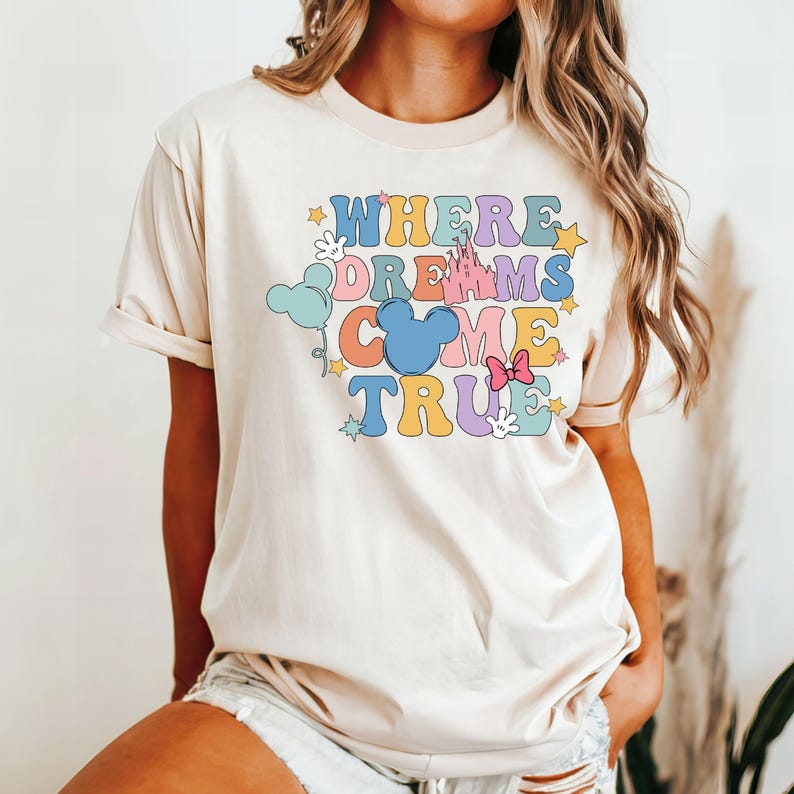 May include: A light beige t-shirt with a colorful graphic that says "Where Dreams Come True" with a Mickey Mouse head, a castle, stars, and balloons.