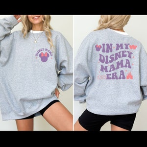 May include: Gray sweatshirt with a purple and pink graphic on the front that says "Disney Mama" with a Mickey Mouse head. The back of the sweatshirt has a purple and pink graphic that says "In My Disney Mama Era" with a Mickey Mouse head and a castle.