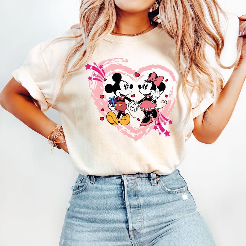 May include: Cream-colored t-shirt featuring a graphic of Mickey and Minnie Mouse holding hands inside a pink heart. Mickey Mouse is holding a gift. Pink stars and hearts surround the heart design.