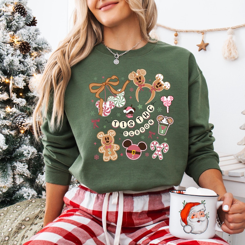 May include: Green sweatshirt with a festive design featuring gingerbread men, Mickey Mouse ears, candy canes, and the phrase "Tis the Season." The design includes pink, white, and brown accents. A white mug with a Santa Claus graphic is held in the hand.