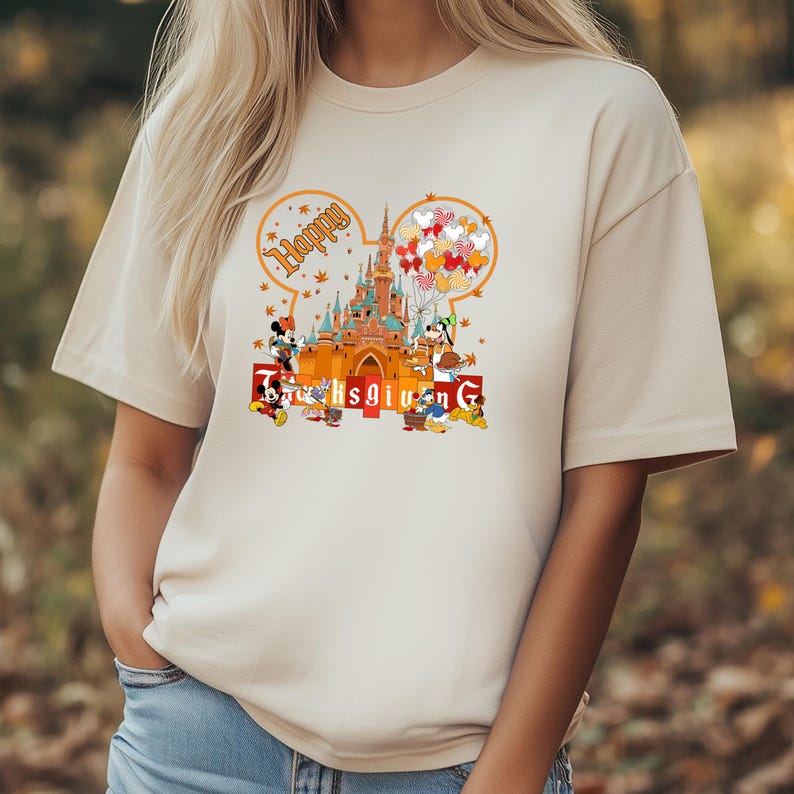 May include: Beige t-shirt featuring a colorful graphic of a castle, Mickey Mouse, Minnie Mouse, Donald Duck, and Goofy. The design includes the words "Happy Thanksgiving" in a festive font, with Mickey Mouse ears and fall leaves.