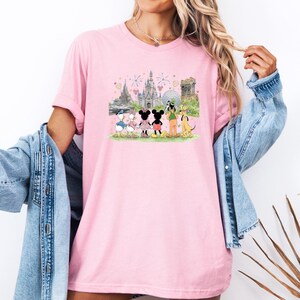 May include: Pink t-shirt featuring a colorful illustration of Disney characters and landmarks. The design includes Mickey and Minnie Mouse, Donald and Daisy Duck, Goofy, and Pluto, with Cinderella's castle and fireworks in the background.