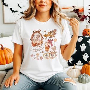 Puede incluir: Camiseta blanca con un gr&aacute;fico de tem&aacute;tica oto&ntilde;al con un castillo, calabazas y personajes de dibujos animados. El dise&ntilde;o incluye hojas de oto&ntilde;o y un marco en forma de coraz&oacute;n de Mickey y Minnie Mouse. La camiseta es de cuello redondo y manga corta.