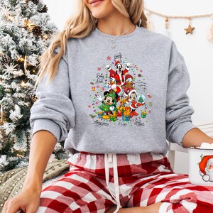 K&ouml;nnte beinhalten: Ein graues Weihnachts-Sweatshirt mit Disney-Figuren in Form eines Weihnachtsbaums. Mickey, Minnie, Goofy, Donald und Pluto sind abgebildet, zusammen mit den Worten "Merry Christmas" und den Namen der Charaktere.