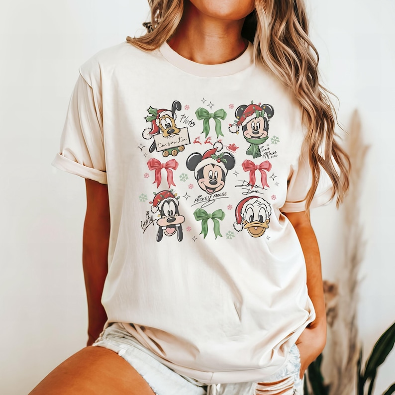 May include: Beige t-shirt featuring cartoon characters Mickey Mouse, Minnie Mouse, Donald Duck, and Goofy in Santa hats and Christmas bows. The shirt has the text "to Santa" and character signatures. Festive holiday apparel.