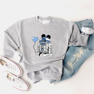 May include: A gray sweatshirt featuring a graphic of R2-D2 with Mickey Mouse ears and a blue heart-shaped balloon. The design is in blue, white, and black. The sweatshirt is paired with blue jeans and white sneakers.