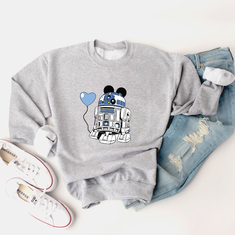 May include: A gray sweatshirt featuring a graphic of R2-D2 with Mickey Mouse ears and a blue heart-shaped balloon. The design is in blue, white, and black. The sweatshirt is paired with blue jeans and white sneakers.