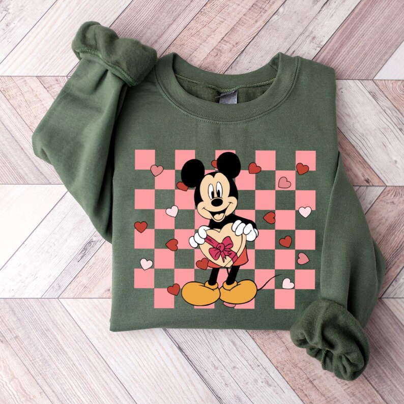 Pode incluir: Camisola verde com um padr&atilde;o xadrez rosa e branco e uma imagem de desenho animado do Mickey Mouse segurando uma caixa de chocolates em forma de cora&ccedil;&atilde;o.