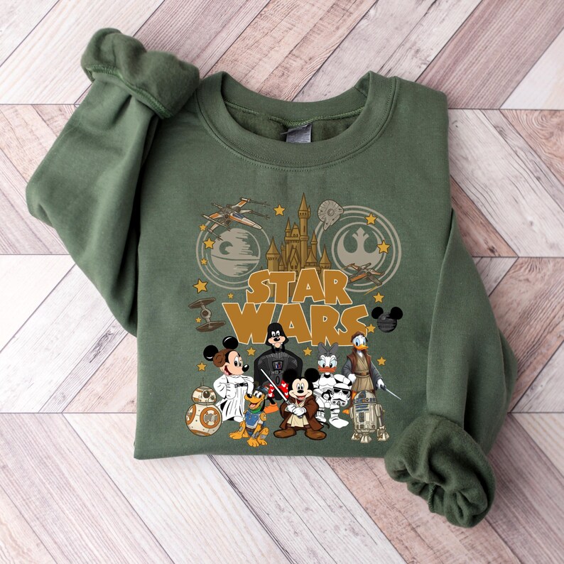 May include: Olive green Star Wars sweatshirt featuring Mickey Mouse, Darth Vader, and other characters. The design includes the Star Wars logo, a castle, and various space vehicles. The sleeves are folded, and the sweatshirt is displayed on a wooden surface.