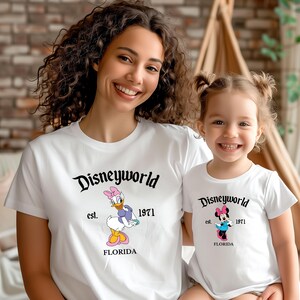 May include: White t-shirts with "Disneyworld" printed in black script. One shirt features Daisy Duck, the other Minnie Mouse, both with "est. 1971 FLORIDA". The shirts are worn by a woman and a child.
