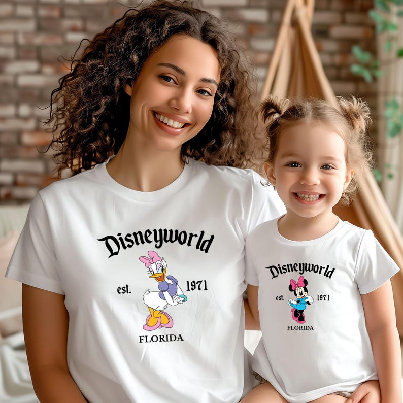 May include: White t-shirts with "Disneyworld" printed in black script. One shirt features Daisy Duck, the other Minnie Mouse, both with "est. 1971 FLORIDA". The shirts are worn by a woman and a child.