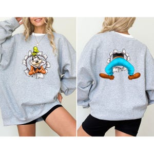 K&ouml;nnte beinhalten: Ein hellgraues Sweatshirt mit einem Cartoon-Charakter-Design. Die Vorderseite des Sweatshirts zeigt das Gesicht der Figur, das aus einem zerrissenen Loch herausragt. Die R&uuml;ckseite des Sweatshirts zeigt die Beine und F&uuml;&szlig;e der Figur.