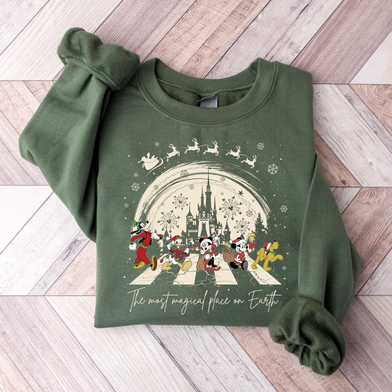 May include: A green sweatshirt with a Christmas-themed design featuring Mickey Mouse and friends walking across a street. The design includes a castle, fireworks, and snowflakes. The text on the sweatshirt reads 'The most magical place on Earth.'