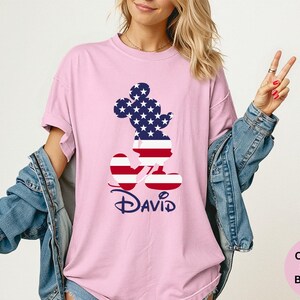 May include: A light pink t-shirt featuring a patriotic Mickey Mouse design. The Mickey Mouse silhouette is filled with the American flag pattern. The name "David" is written in a navy blue script font below the design.